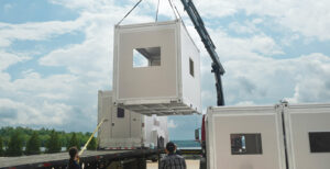 Modular structure unloading. Four structures, one on a crane, one on a truck, two on the ground.